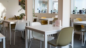 A modern dining area with white tables and chairs, large windows with views of buildings, and decorative items on the windowsills. - Hotell Stortorget (Östersund)