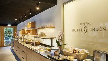 A breakfast buffet area with various food items, a toaster, and a view of the hotel name 'ELZLAND HOTEL O LINDEN'.