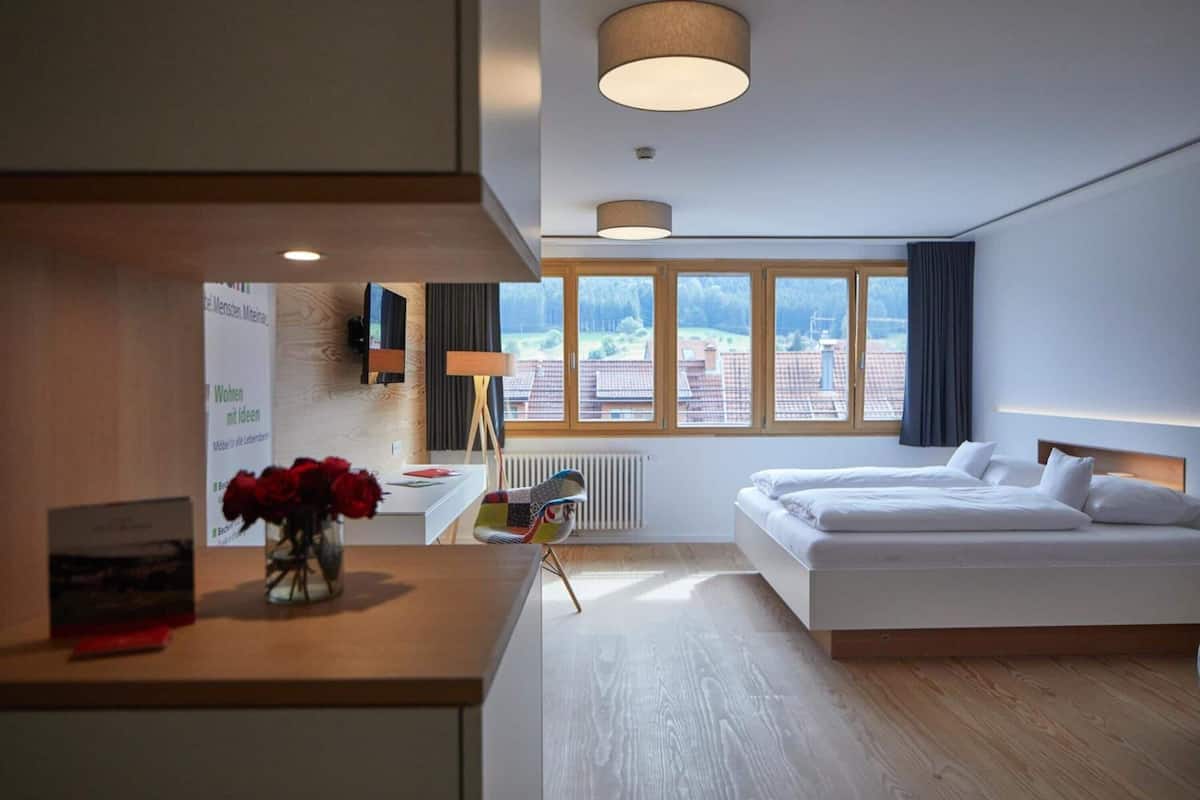 A hotel room with a bed, a desk, a chair, a window with a view, and a vase with flowers.