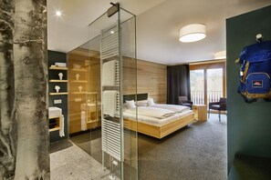 Ein modernes Hotelzimmer mit Glastrennwand, einem Holzbett, einem kleinen Schreibtisch und Blick ins Freie.