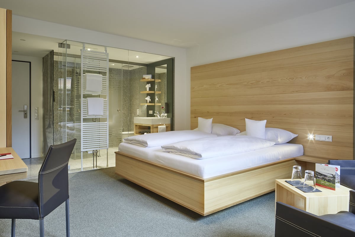 A modern hotel room with a large bed, a desk with a chair, a glass shower enclosure, and a wooden headboard.