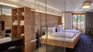Una habitación de hotel moderna con separador de vidrio, armario de madera, cama con ropa blanca, baño con espejo y balcón con vistas a la ciudad.