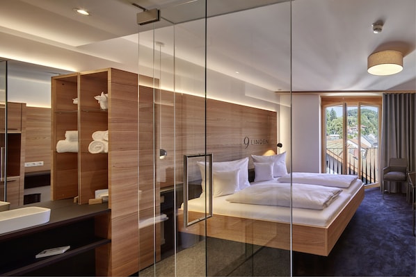 A modern hotel room with a glass partition, a wooden wardrobe, a bed with white linens, a bathroom with a mirror, and a balcony with a view of the city.