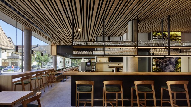 Un área de bar moderna con sillas y mesas de madera, una barra con estantes llenos de vasos y vistas a la calle.