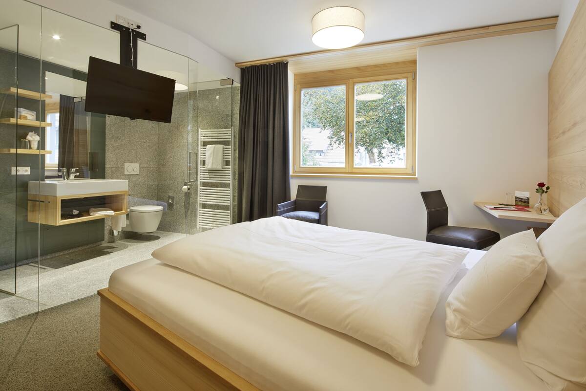 A modern hotel room with a large bed, a glass shower enclosure, a mounted TV, and a window with a view of trees.