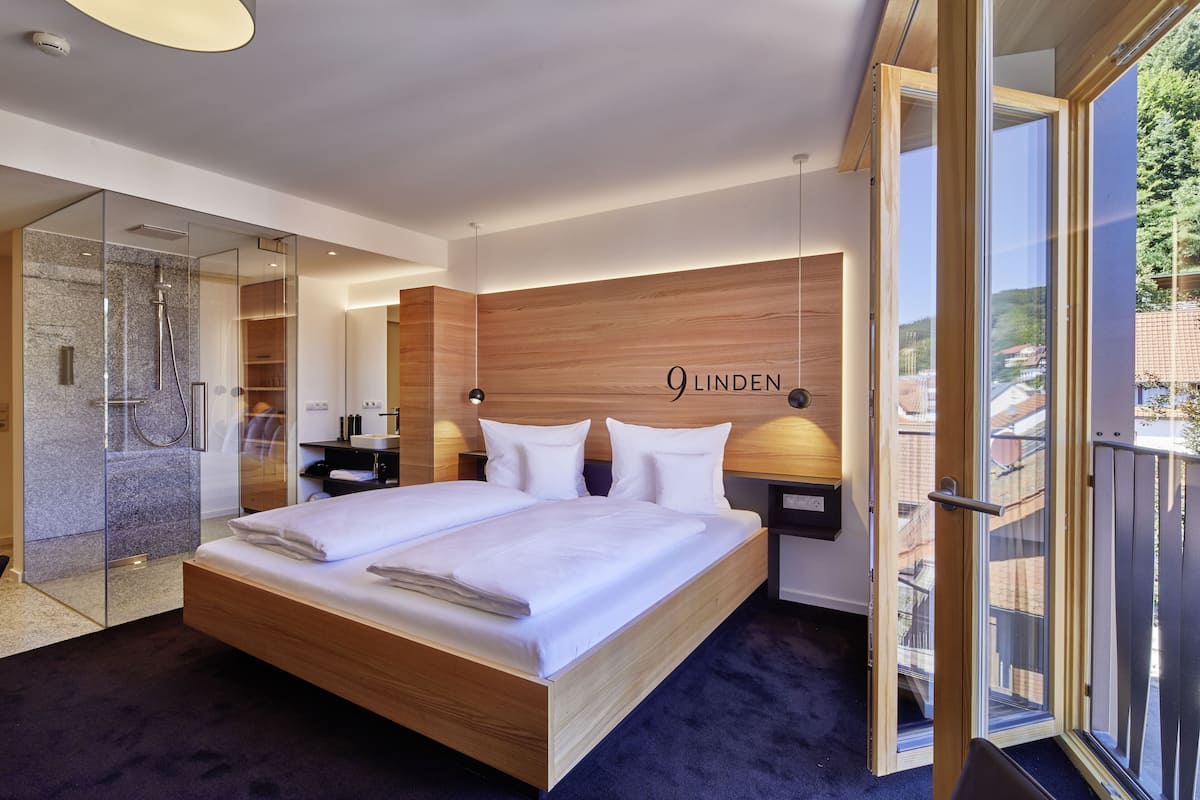 A modern hotel room with a large bed, a glass shower enclosure, wooden headboard, and a view of the city through large windows.