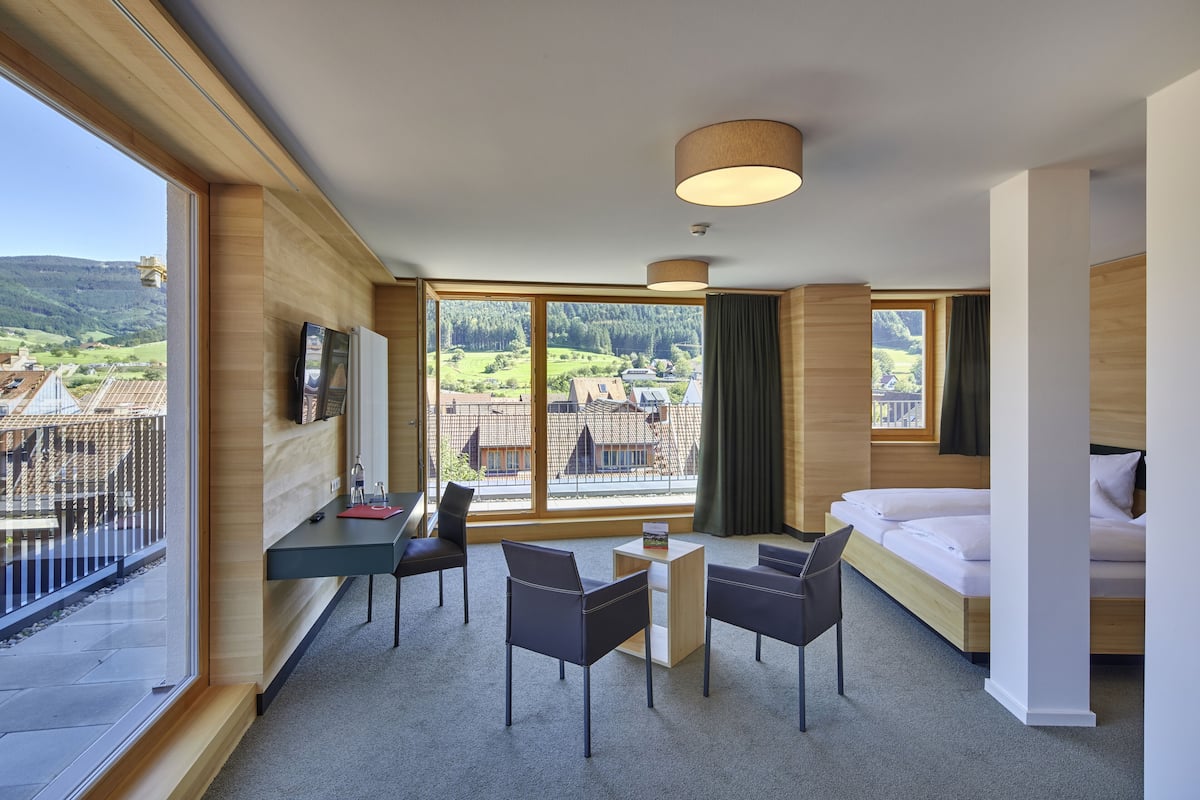 A modern hotel room with a large window offering a view of a village, a bed, a desk with a chair, and a small table.
