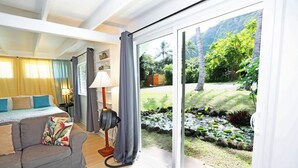 Una habitación con una cama, un sofá gris, una lámpara de pie y vistas a un jardín con un estanque y árboles.