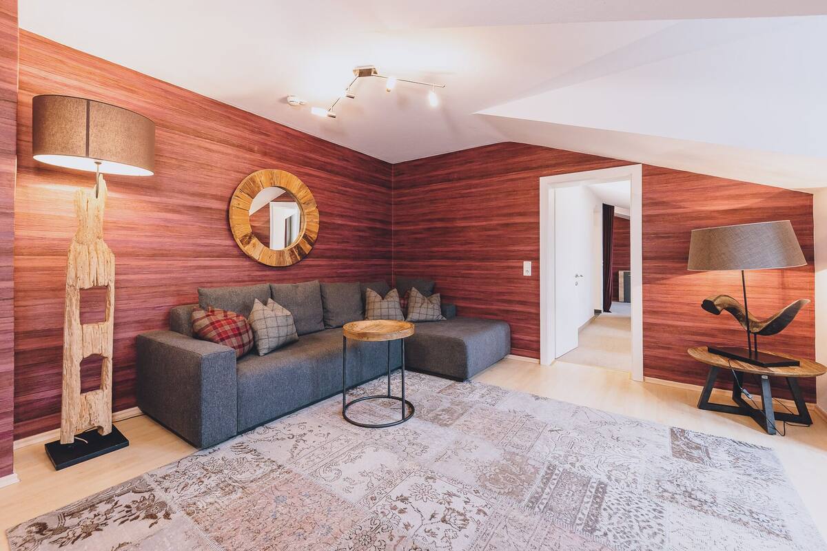 Un salon moderne avec un canapé d’angle gris, un miroir rond, un mur d’accent en bois et un tapis à motifs.