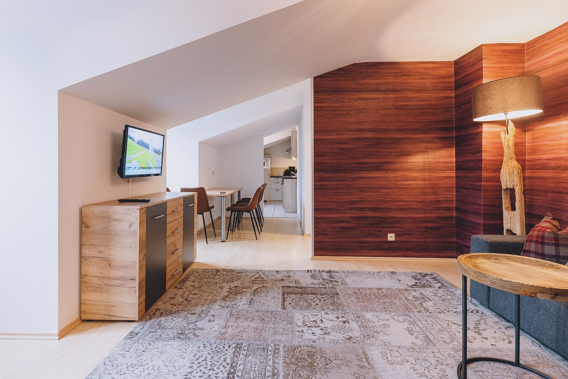 Un salon moderne avec un meuble en bois, un téléviseur, un coin repas avec des chaises, un tapis à motifs et un mur d’accent en bois.