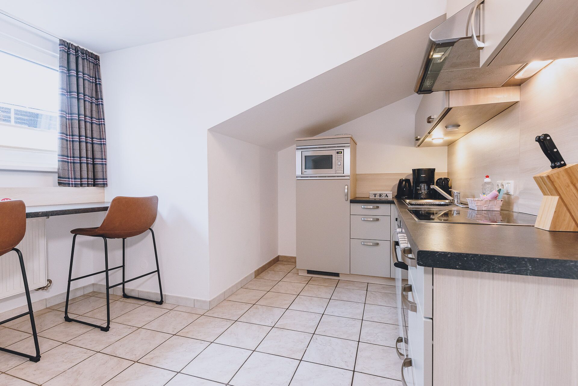 A compact kitchen with a microwave, coffee maker, and a knife block. There are two bar stools and a window with curtains.