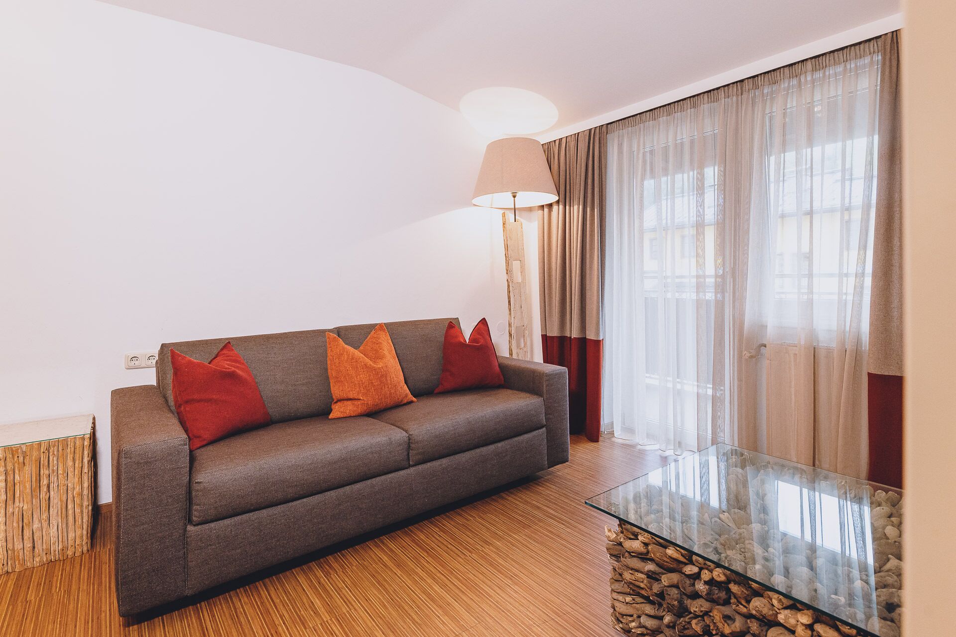 Un salon moderne avec un canapé gris, des coussins rouges et orange, une table basse en verre et une lampe.
