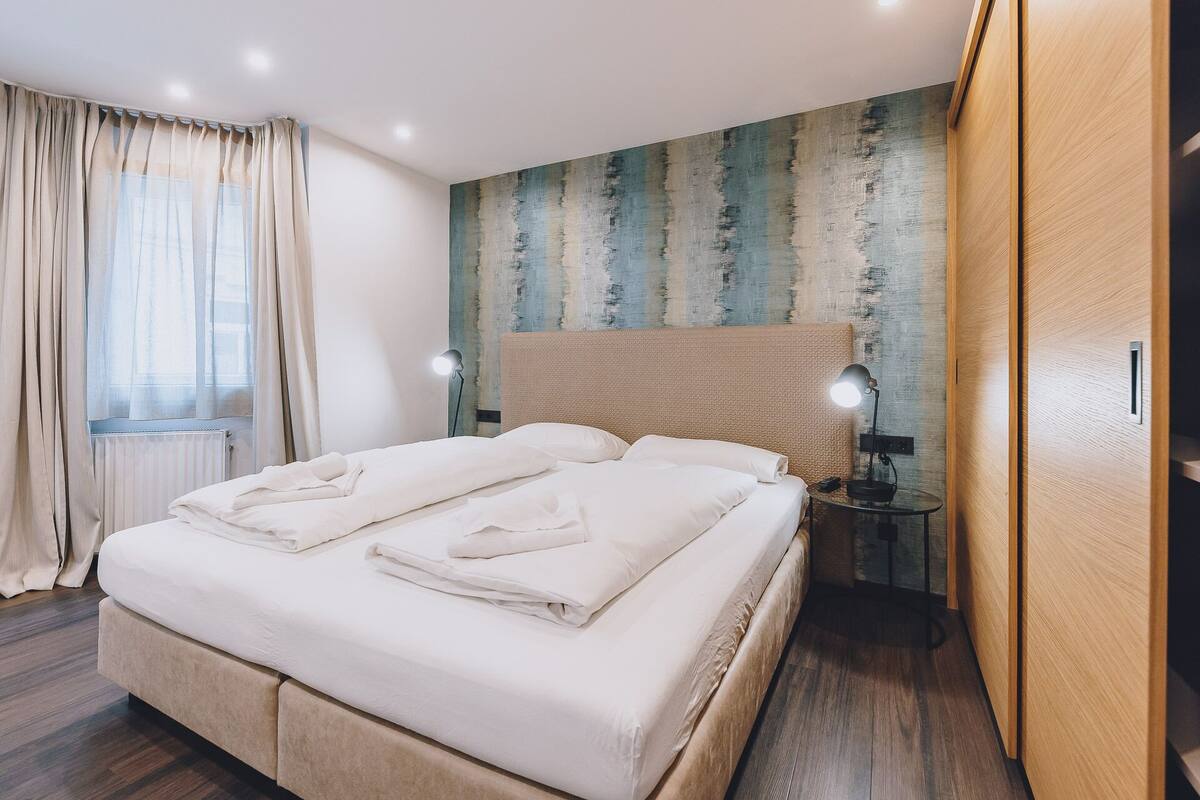 Un lit bien fait, avec du linge de lit et des serviettes blanches, une table de chevet ronde avec une lampe, et un mur texturé derrière le lit.