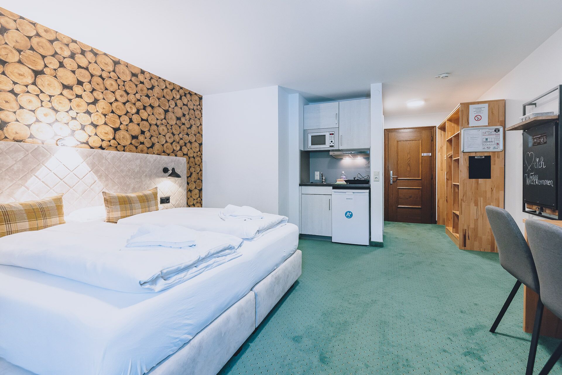 A hotel room with a bed, a kitchenette, a wooden wardrobe, and a desk.