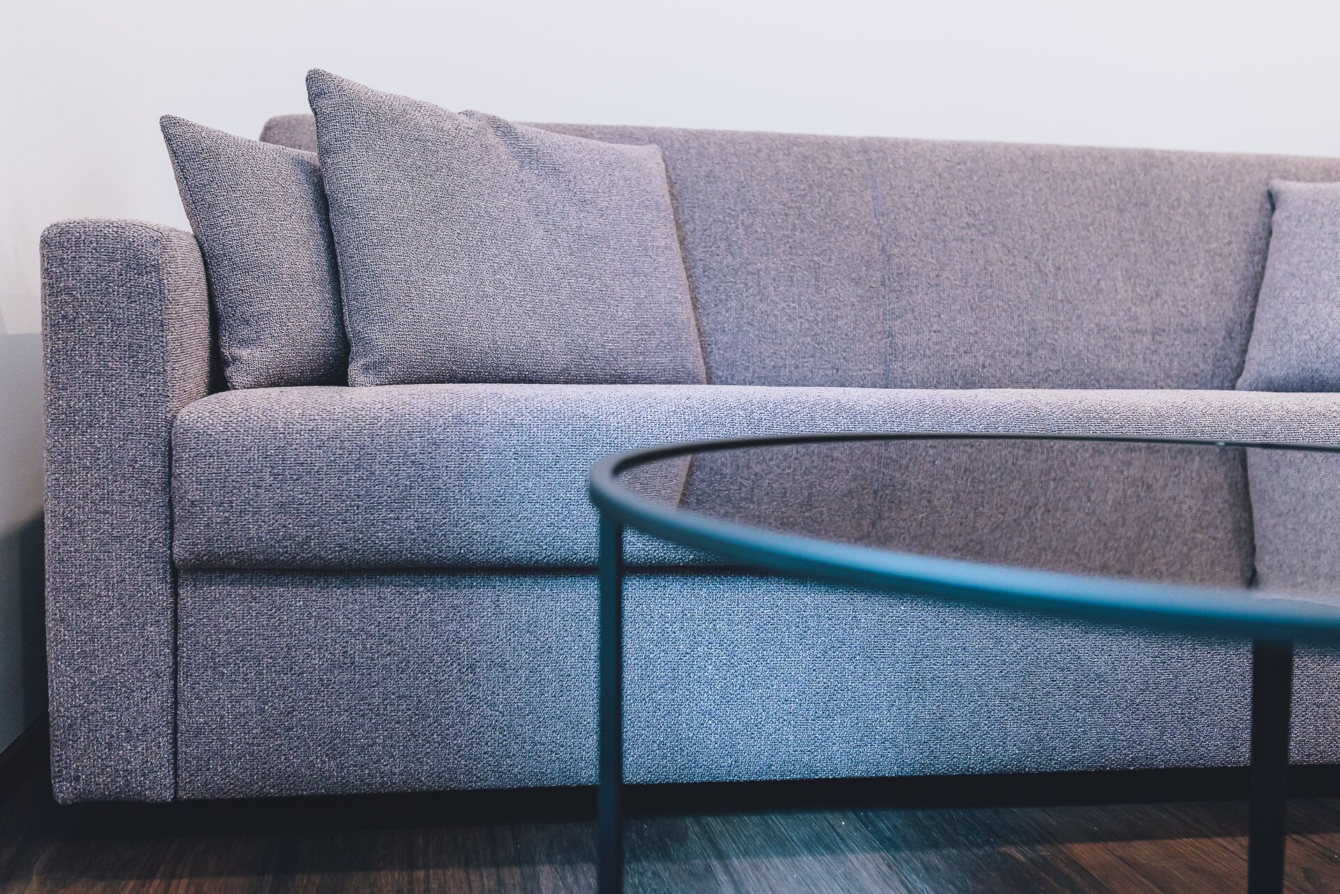 A grey sofa with a textured fabric, a glass coffee table with a metal frame, and a dark wooden floor.