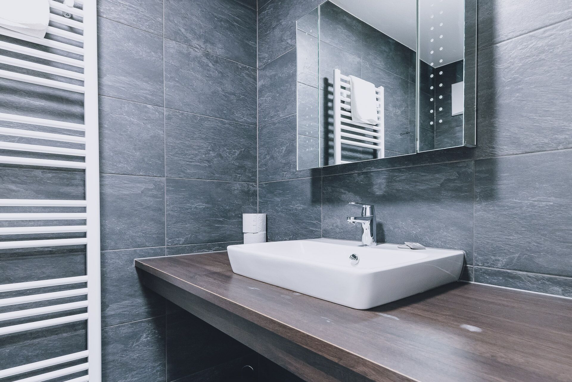 Un bagno moderno con pareti piastrellate in tonalità scura, un lavandino bianco e un piano d'appoggio in legno.