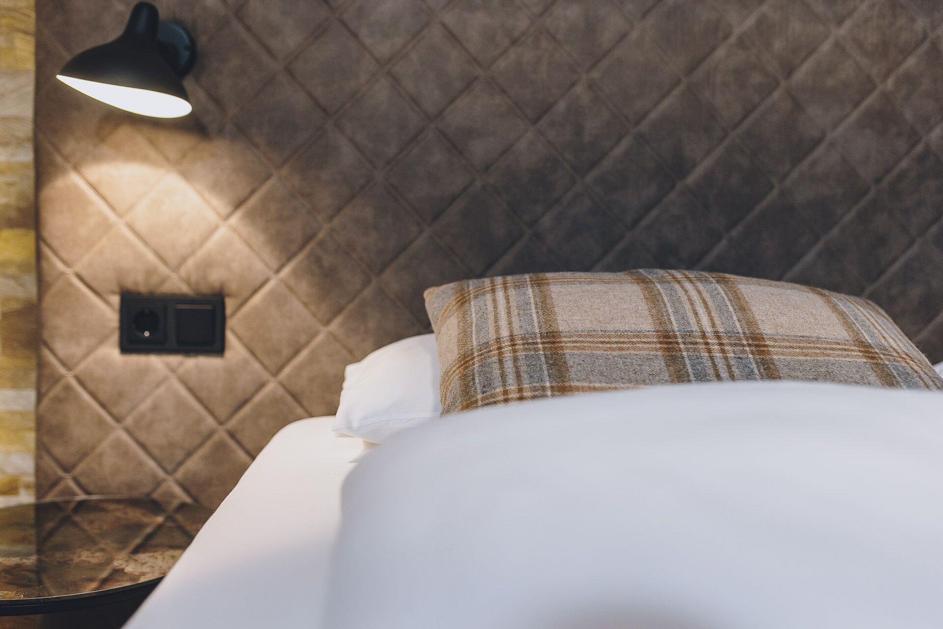 Een bed met een geruit kussen en een wandlamp.