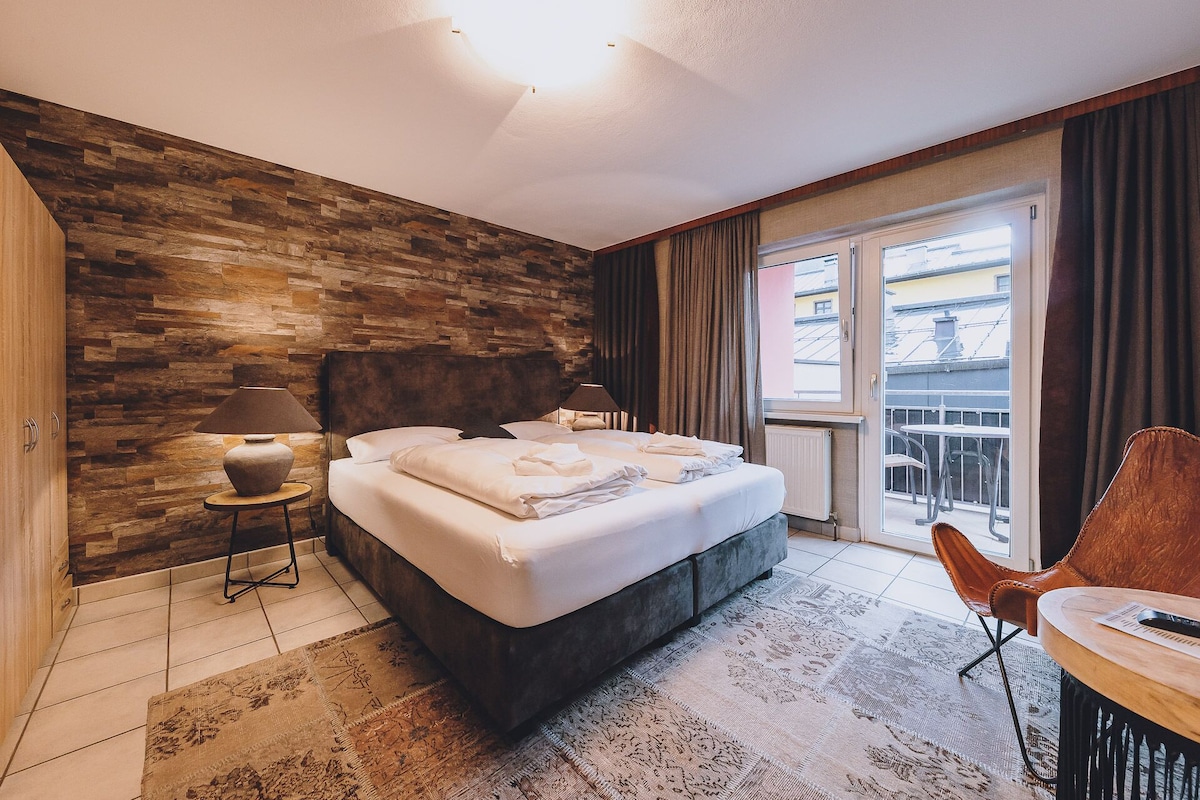 A modern hotel room with a large bed, a wooden accent wall, a balcony with a view, and a small seating area.