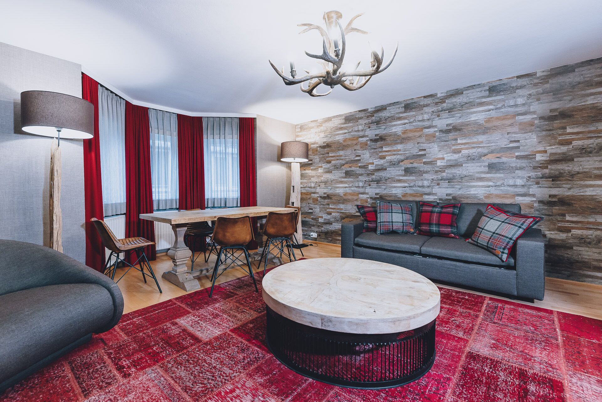 Een moderne woonkamer met een stenen muur, een ronde salontafel, een bank met geruite kussens, een eettafel met stoelen en een kroonluchter.