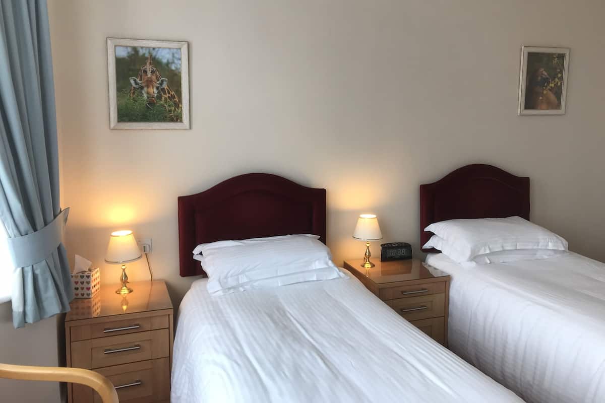 A hotel room with two beds, each with a wooden headboard and white bedding. There are bedside tables with lamps and a digital clock. The room has a window with curtains, and two framed pictures on the wall.