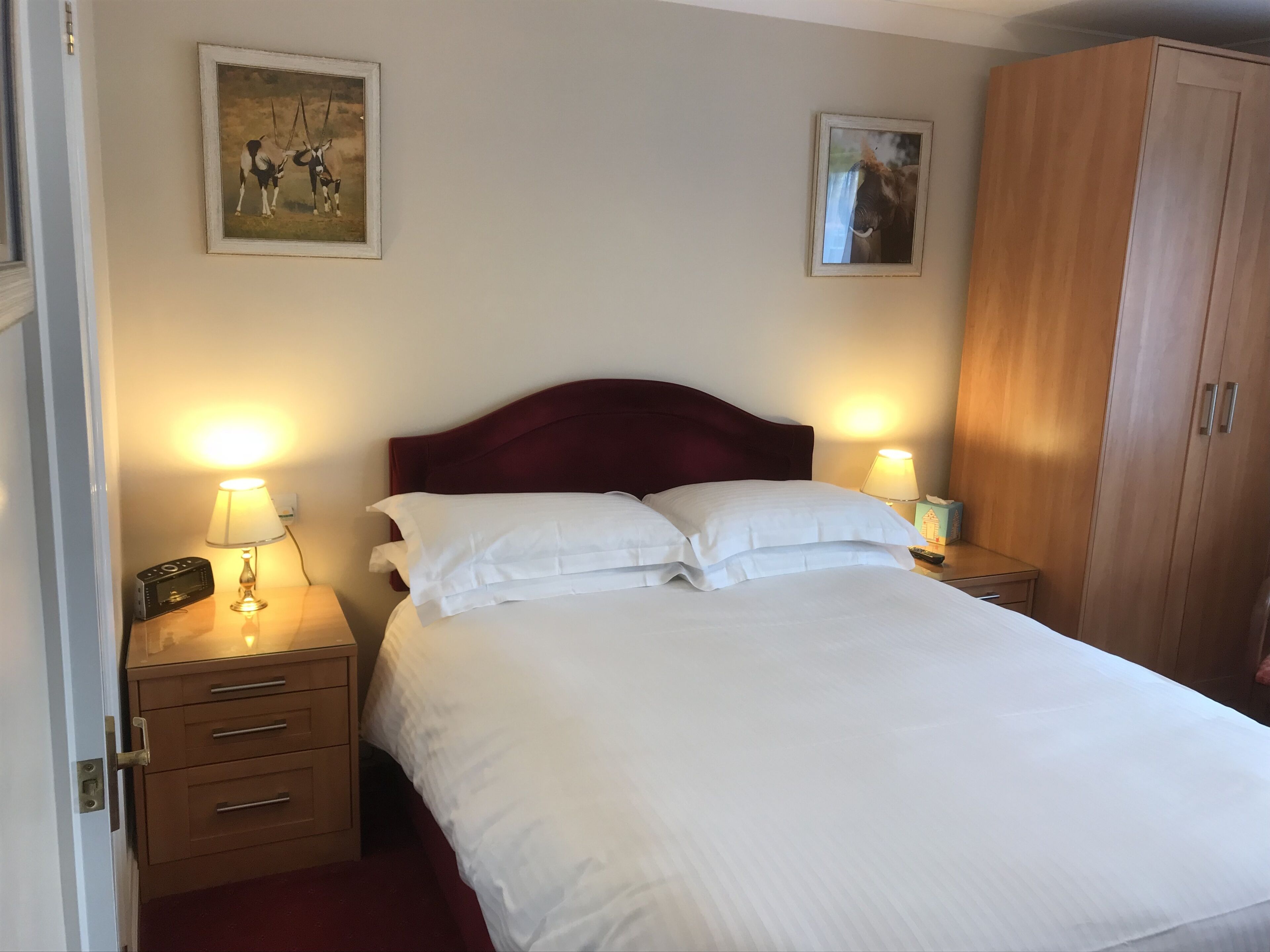 A neatly made bed with white linens, two bedside tables with lamps, a wooden wardrobe, and two framed pictures on the walls.