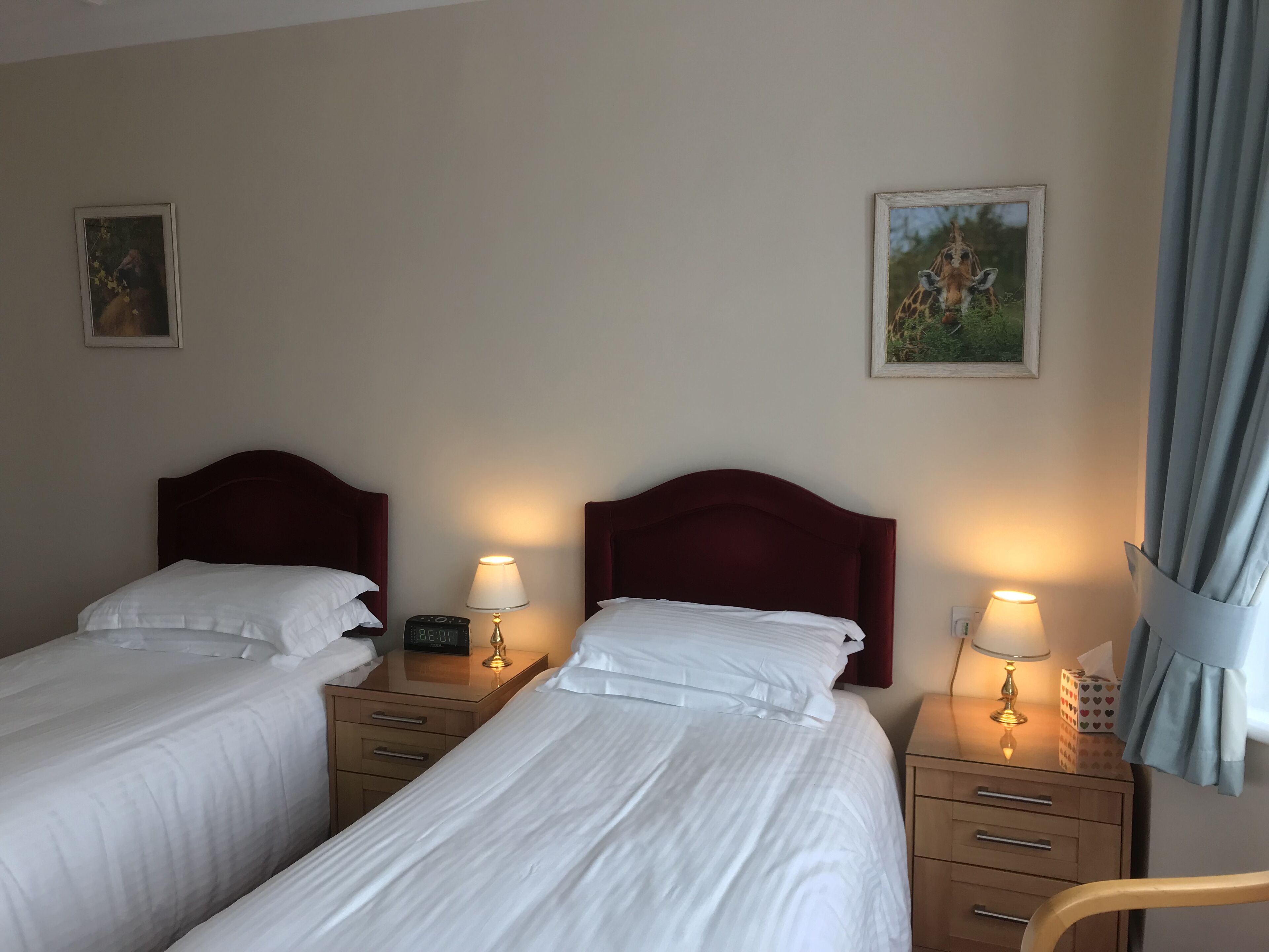 Ein Hotelzimmer mit zwei Einzelbetten, Holz-Nachttischen, Nachttischlampen, einer Uhr und zwei gerahmten Bildern an der Wand.