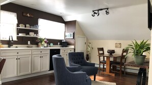 A modern kitchen with white cabinets, a dining area with a wooden table and chairs, and a living space with a blue armchair and a white rug.
