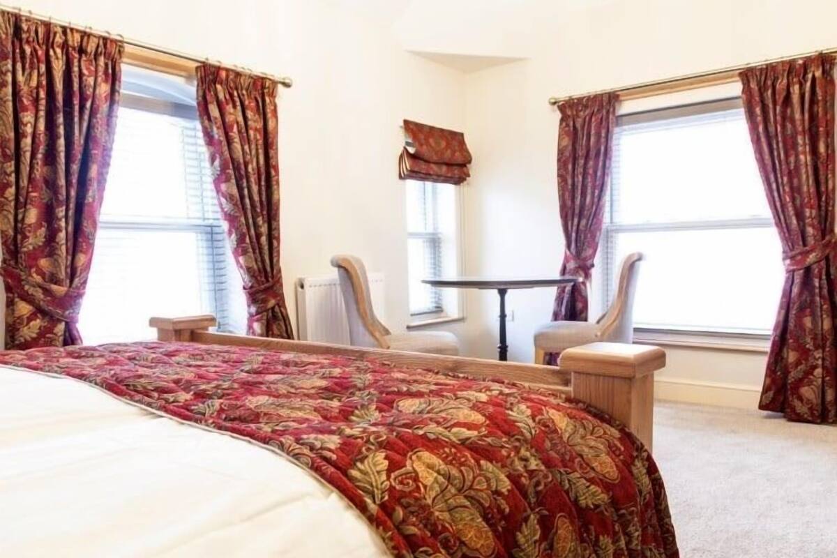 Habitació d'hotel amb un llit gran, dues cadires, una taula petita i una finestra amb cortines.