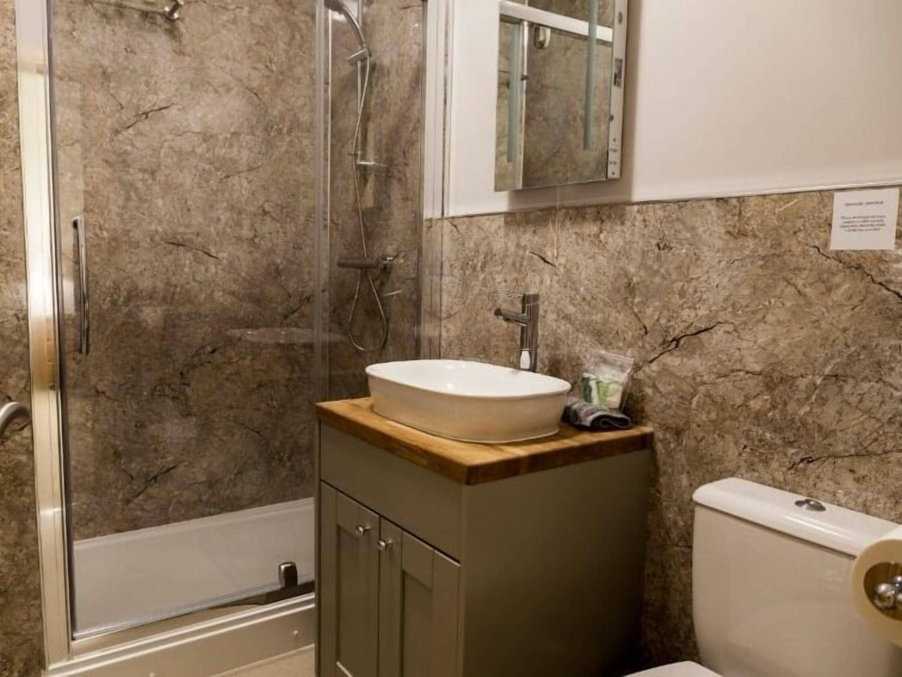 Un baño con ducha de vidrio, un lavamanos blanco sobre encimera de madera y un inodoro.
