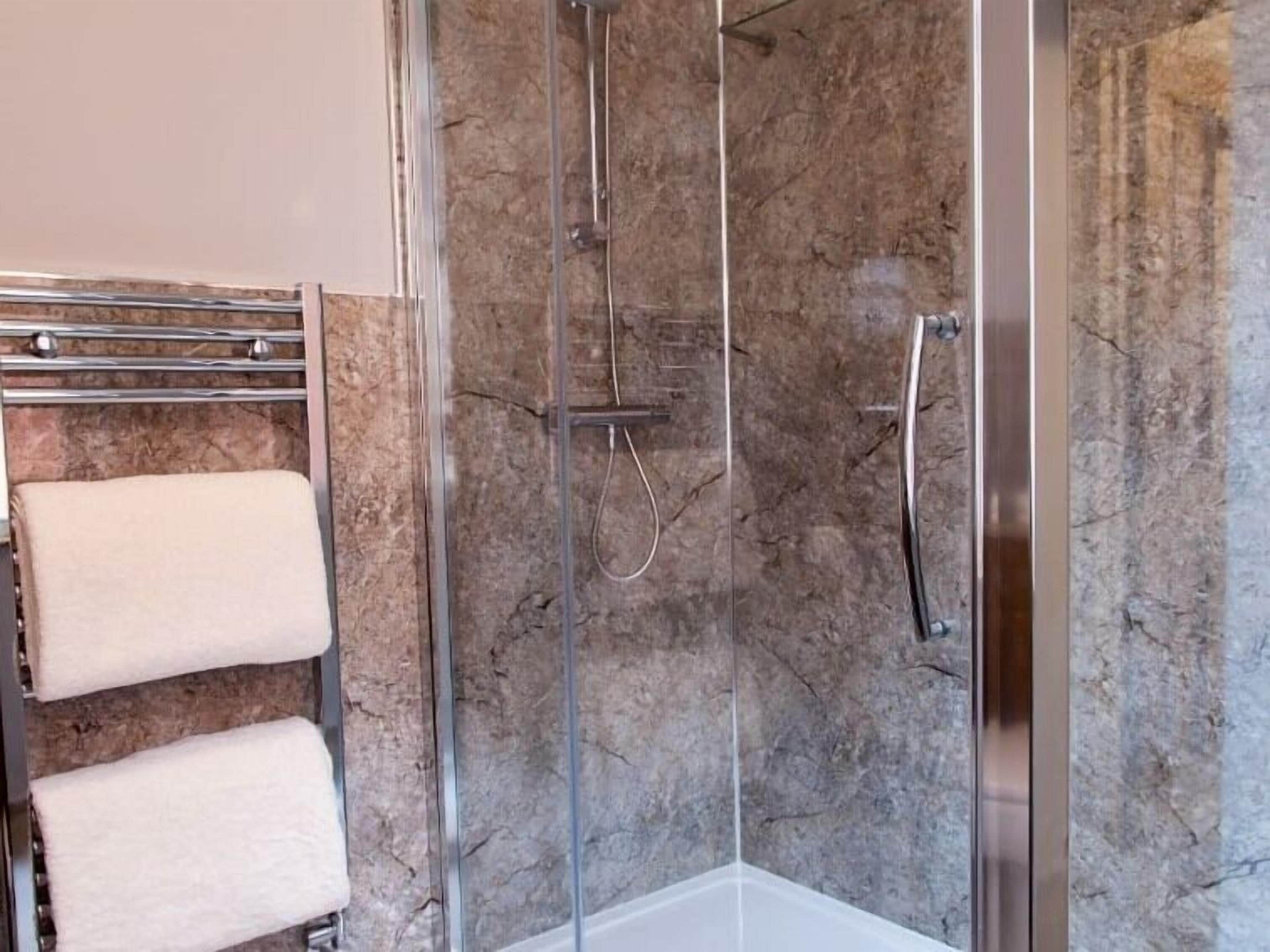A glass-enclosed shower with a marble wall, a towel rack with two towels, and a showerhead.