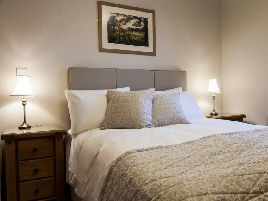 Un letto rifatto con cura, con copriletto decorato, due cuscini e una testiera. Sono presenti due comodini con lampade, una piccola cassettiera in legno e un quadro incorniciato appeso al muro.