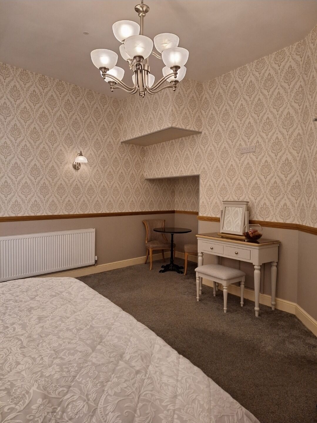 A hotel room with a bed, a desk with a chair, a small table, and a chandelier.