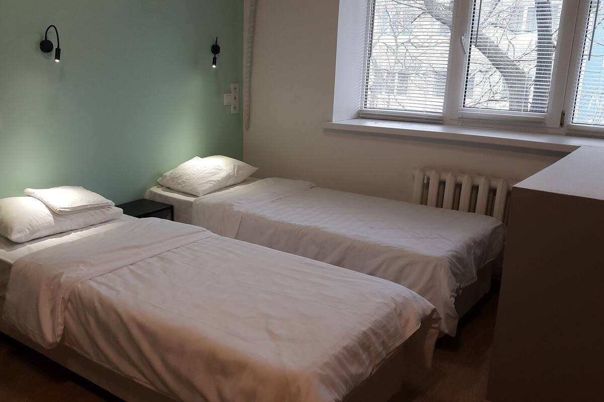 Une chambre avec deux lits simples, une table de chevet, une commode, une fenêtre avec stores et un luminaire fixé au mur.