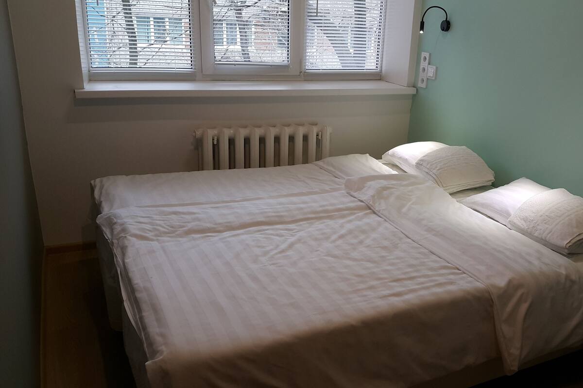 Une chambre avec un lit, un radiateur, une fenêtre avec des stores et une lampe fixée au mur.