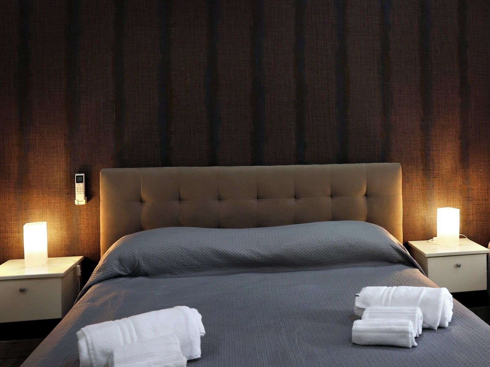 A neatly made bed with a grey bedspread and white towels, two bedside lamps, and a wooden headboard.