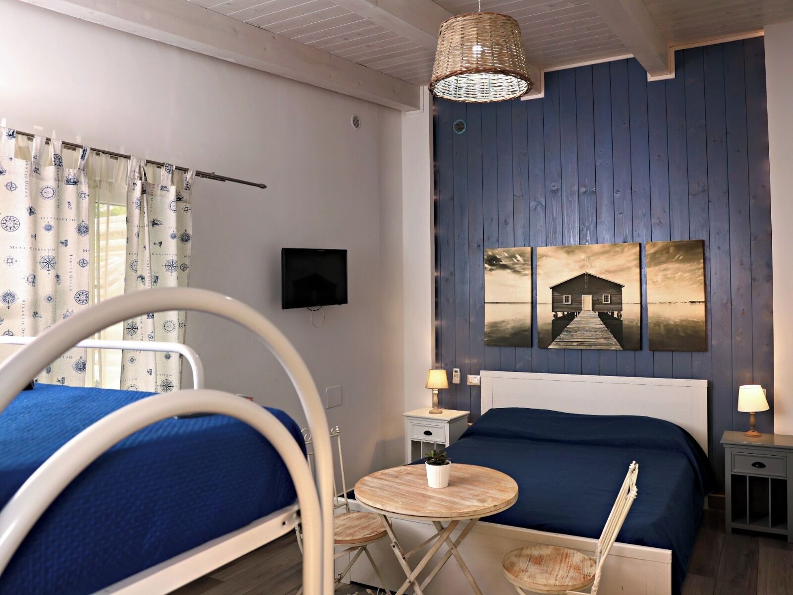 Une chambre à coucher avec un lit, des tables de chevet, une petite table avec une plante, un téléviseur fixé au mur et une grande fenêtre avec des rideaux.