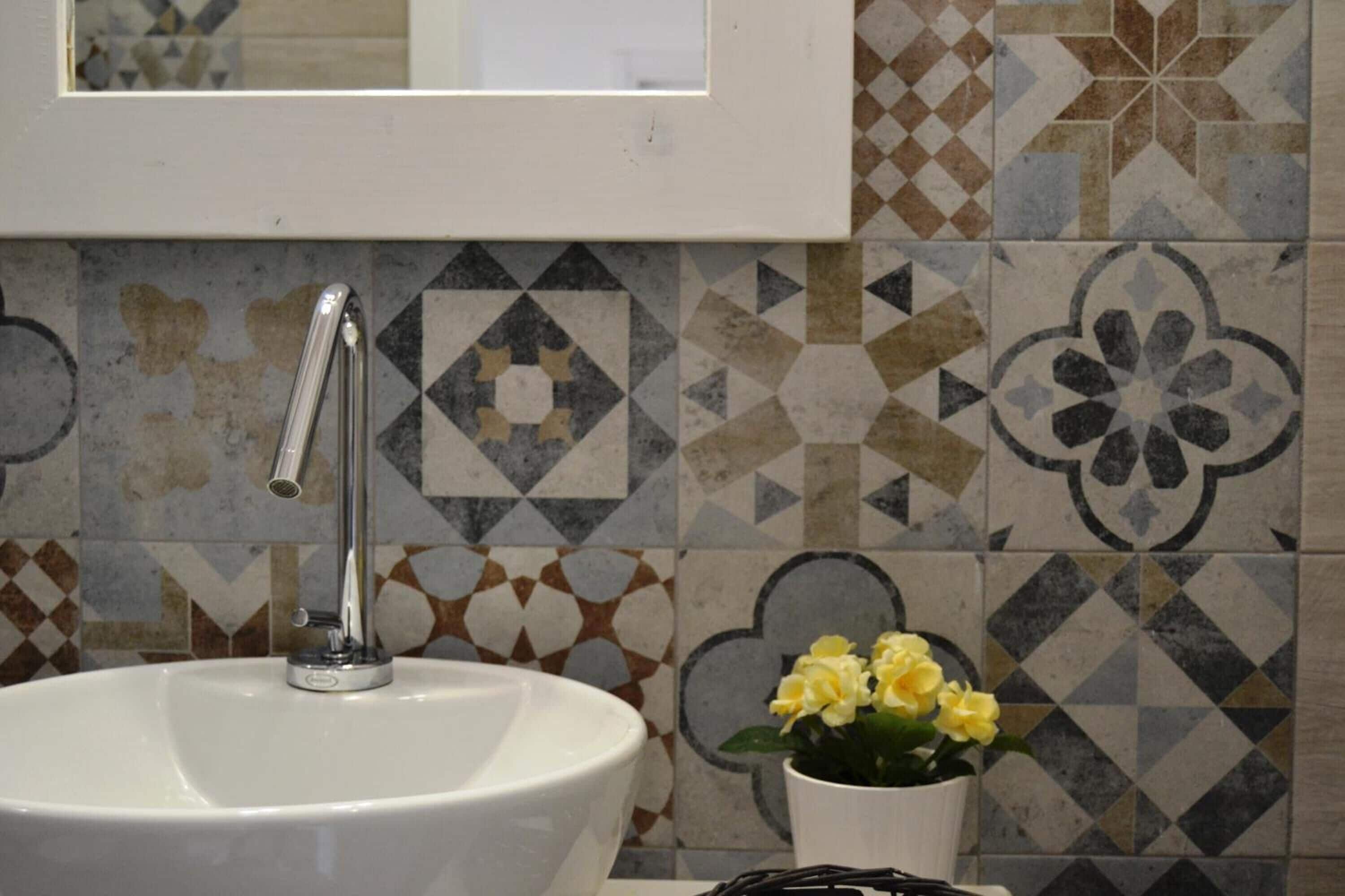 A bathroom with a white sink, a chrome faucet, and a mirror above it. The wall is tiled with geometric patterns in various shades.