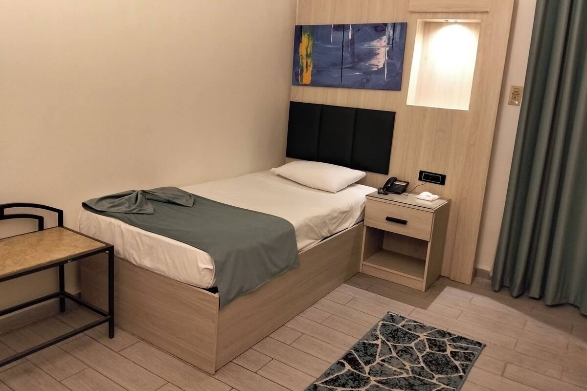 Un lit simple avec une table de chevet, un bureau, une petite table d’appoint, un tableau et une fenêtre avec des rideaux.