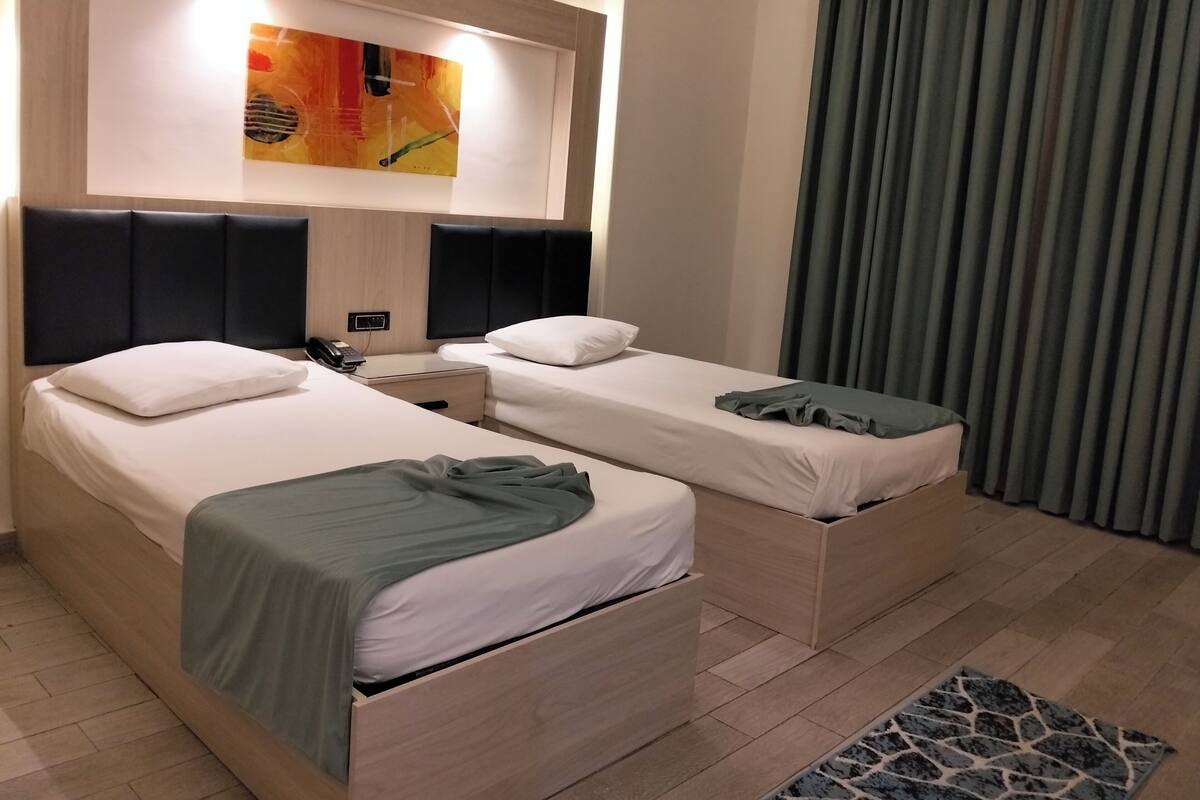 Une chambre d’hôtel avec deux lits, une table de chevet, un téléphone, un tableau au mur et une fenêtre avec des rideaux.