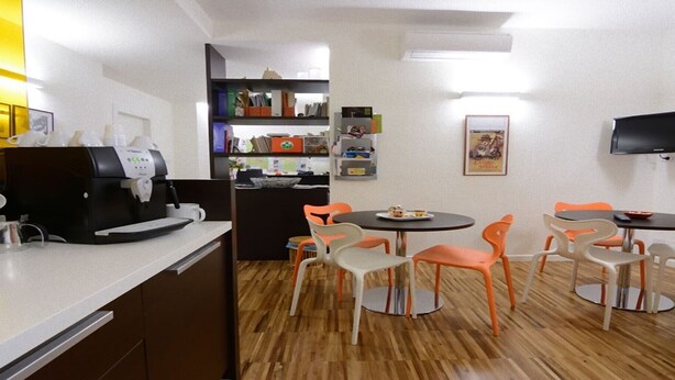 A compact kitchen with a coffee machine, a dining area with orange chairs, and a flat-screen TV mounted on the wall.