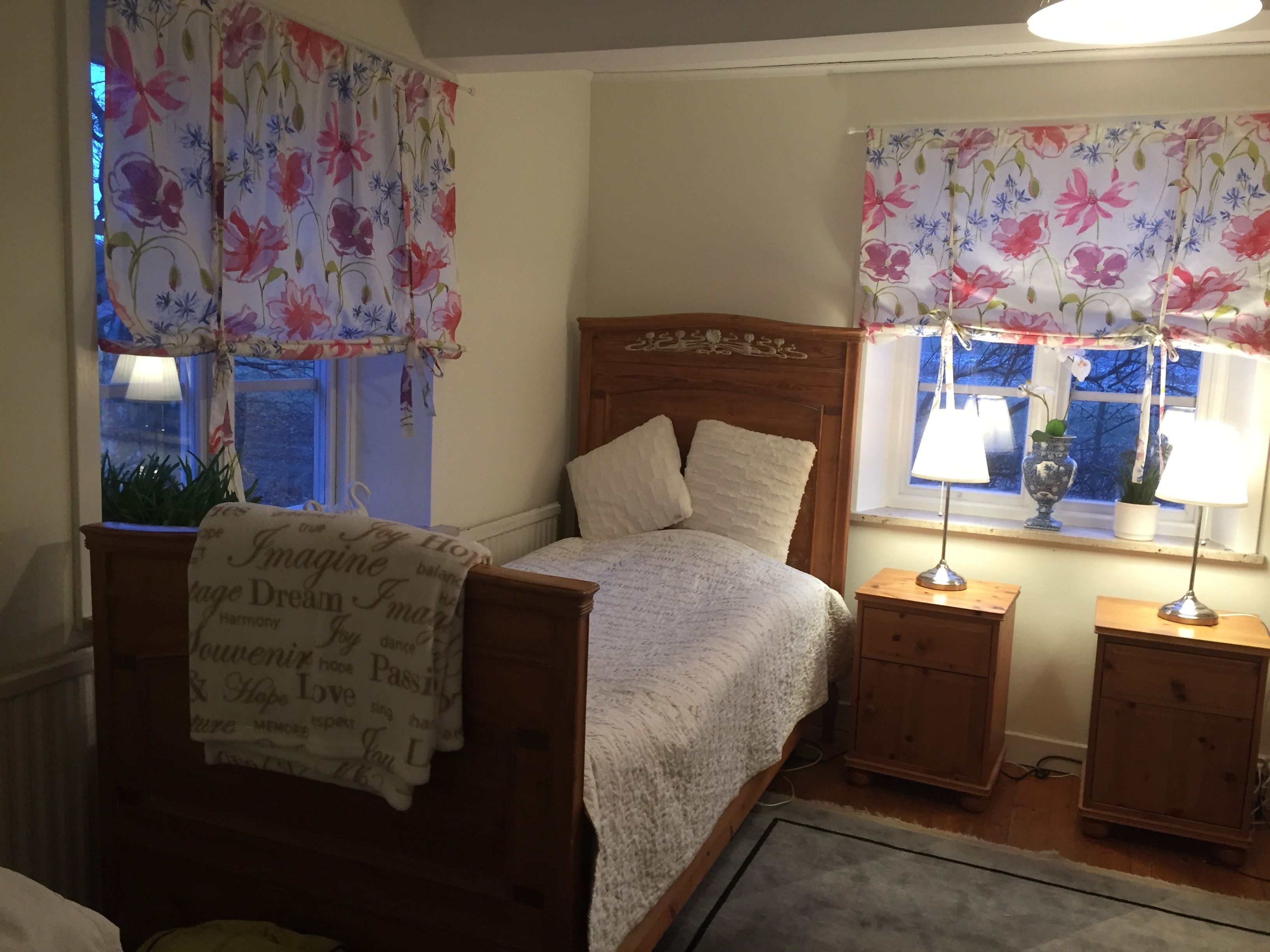 Un dormitorio con una cama de madera, dos mesitas de noche, un ventanal con cortinas florales y un radiador.