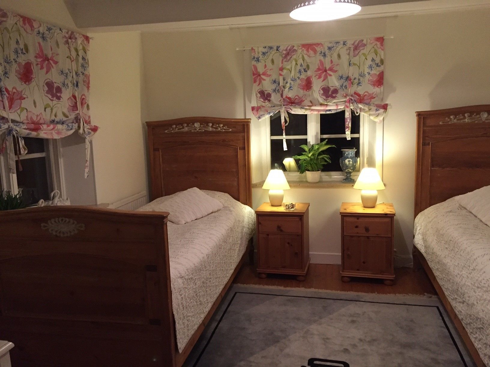 A bedroom with two wooden beds, a window with floral curtains, bedside lamps, and a small potted plant.