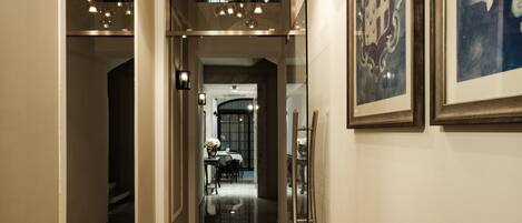 A hallway with a marble floor leading to a dining area with a table and chairs. There are framed artworks on the walls and a chandelier hanging from the ceiling.