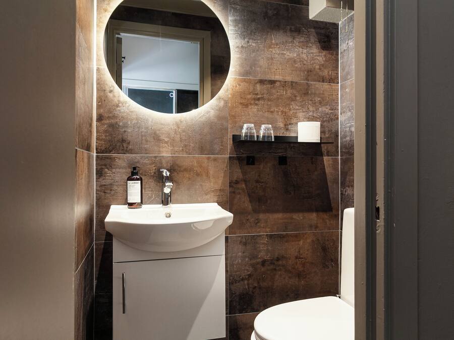 A bathroom with a white sink, a mirror, and a toilet. There is a bottle of shampoo on the sink.