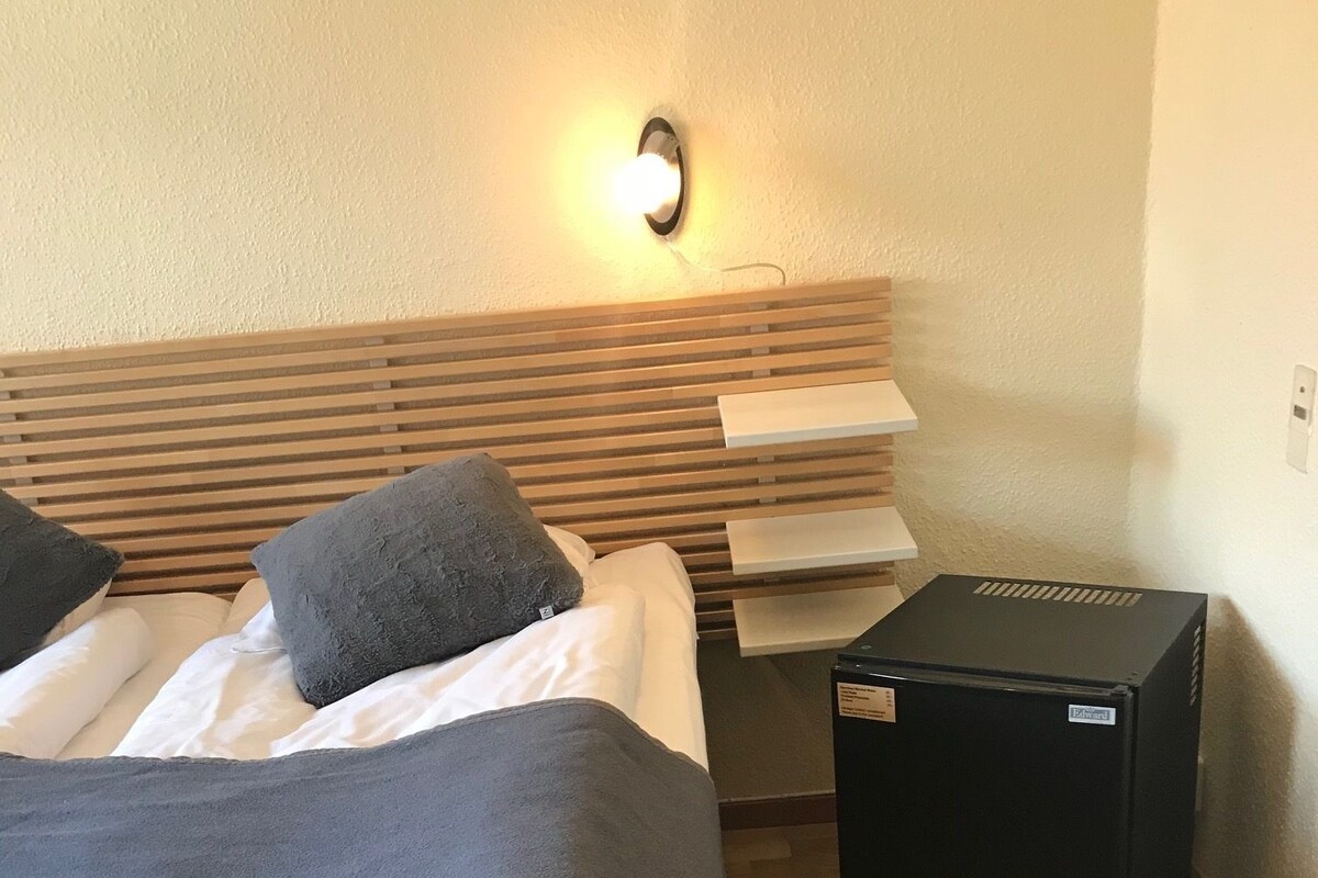 Una habitación de hotel con cama, mesita de noche, lámpara y un altavoz negro.