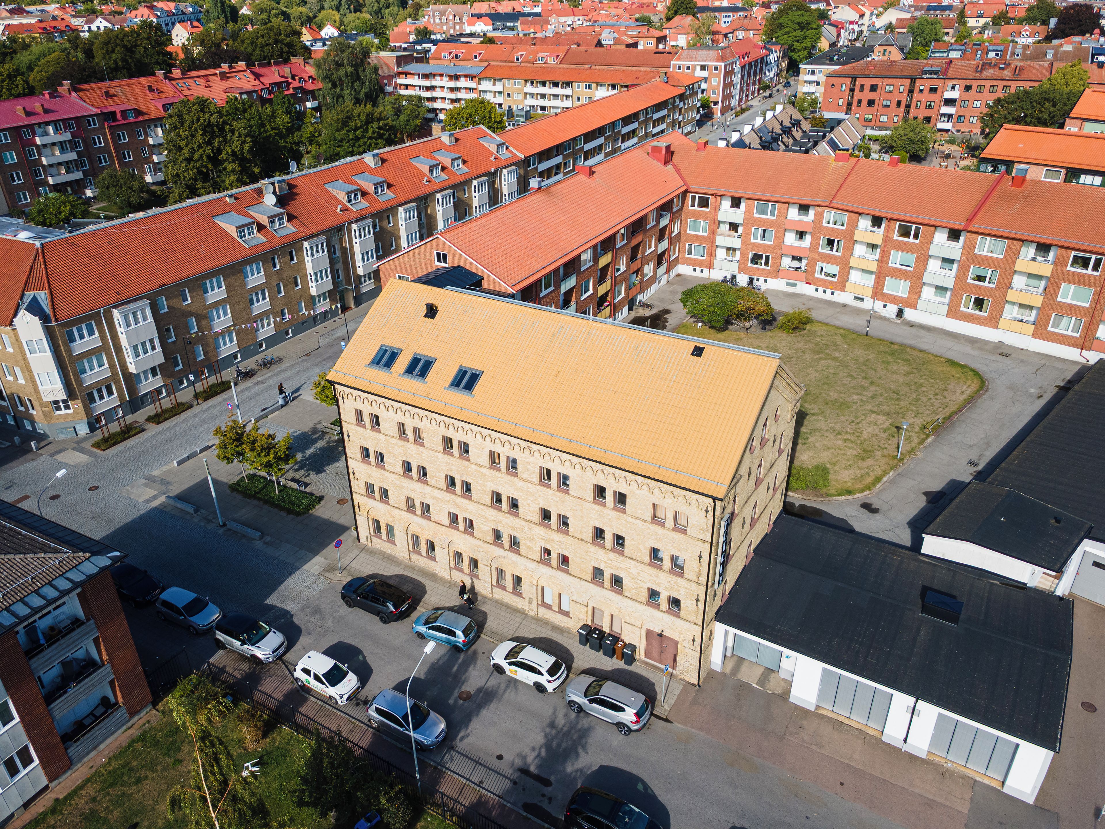 A multi-story building with a flat roof, surrounded by other buildings, a parking lot with cars, and a grassy area.