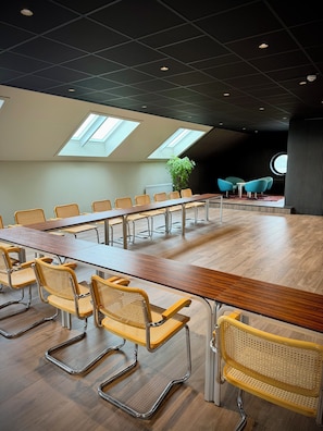 Un salón de conferencias con una larga mesa, varias sillas, una planta en maceta y una mesa redonda con sillas al fondo.