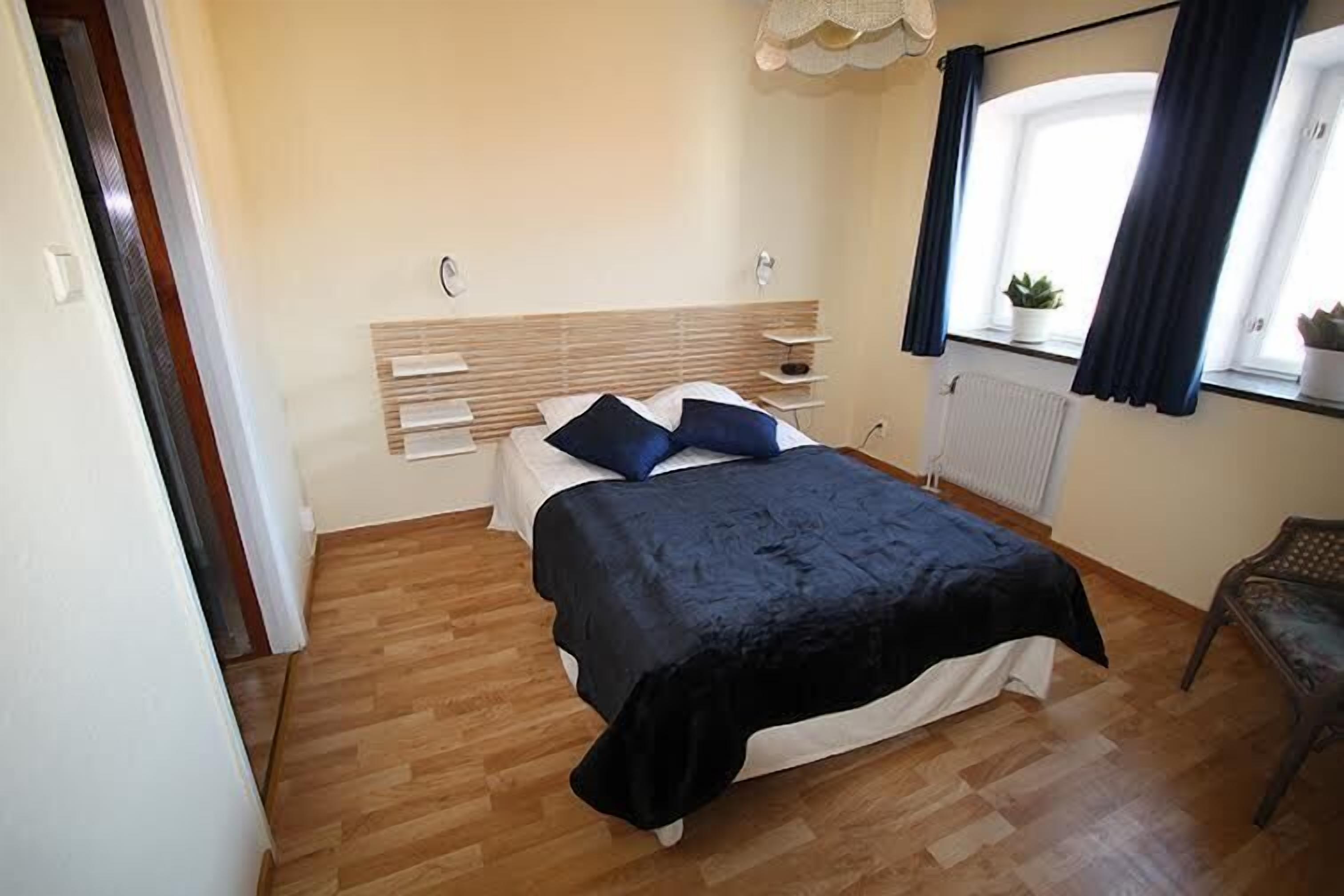 Un dormitorio con cama, piso de madera, una ventana con cortinas y una mesita con una planta.