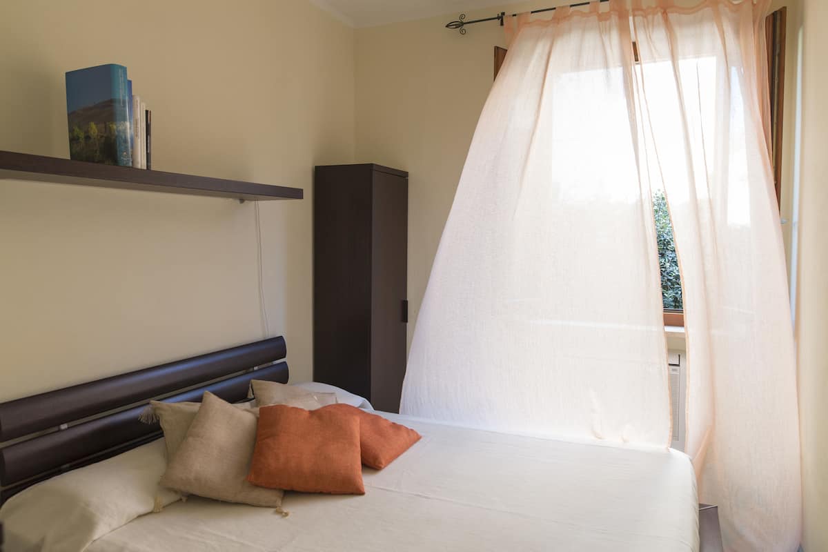 Ein Schlafzimmer mit Bett, Kissen, Nachttisch, Bücherregal und einem Fenster mit leichtem Vorhang.