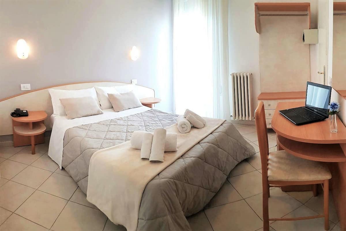 Habitación de hotel con cama, escritorio con una computadora portátil, silla y una mesita de noche con lámpara.